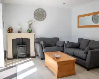 A cozy living room featuring two gray sofas facing a wooden coffee table. A fireplace with a black stove is against the wall, and a large clock hangs above it. A mirror is mounted adjacent to the fireplace, and light streams in from a nearby window.