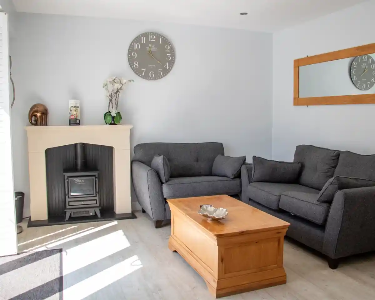 A cozy living room featuring two gray sofas facing a wooden coffee table. A fireplace with a black stove is against the wall, and a large clock hangs above it. A mirror is mounted adjacent to the fireplace, and light streams in from a nearby window.