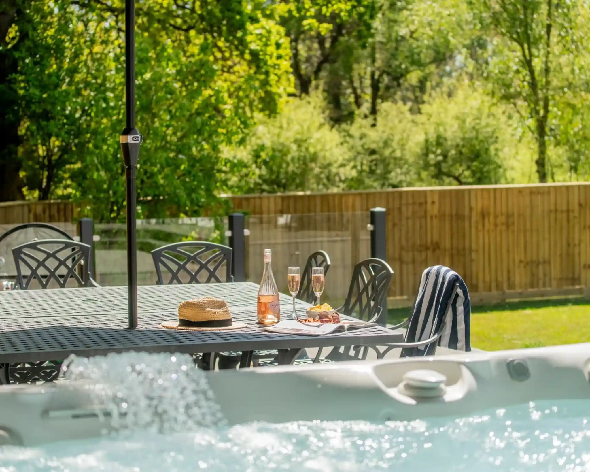 A sunlit outdoor space featuring a dining table with a bottle of rosé, two glasses of sparkling wine, and snacks. Nearby, a hot tub bubbles in the foreground, surrounded by greenery and a wooden fence in the background.