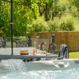 A sunlit outdoor space featuring a dining table with a bottle of rosé, two glasses of sparkling wine, and snacks. Nearby, a hot tub bubbles in the foreground, surrounded by greenery and a wooden fence in the background.