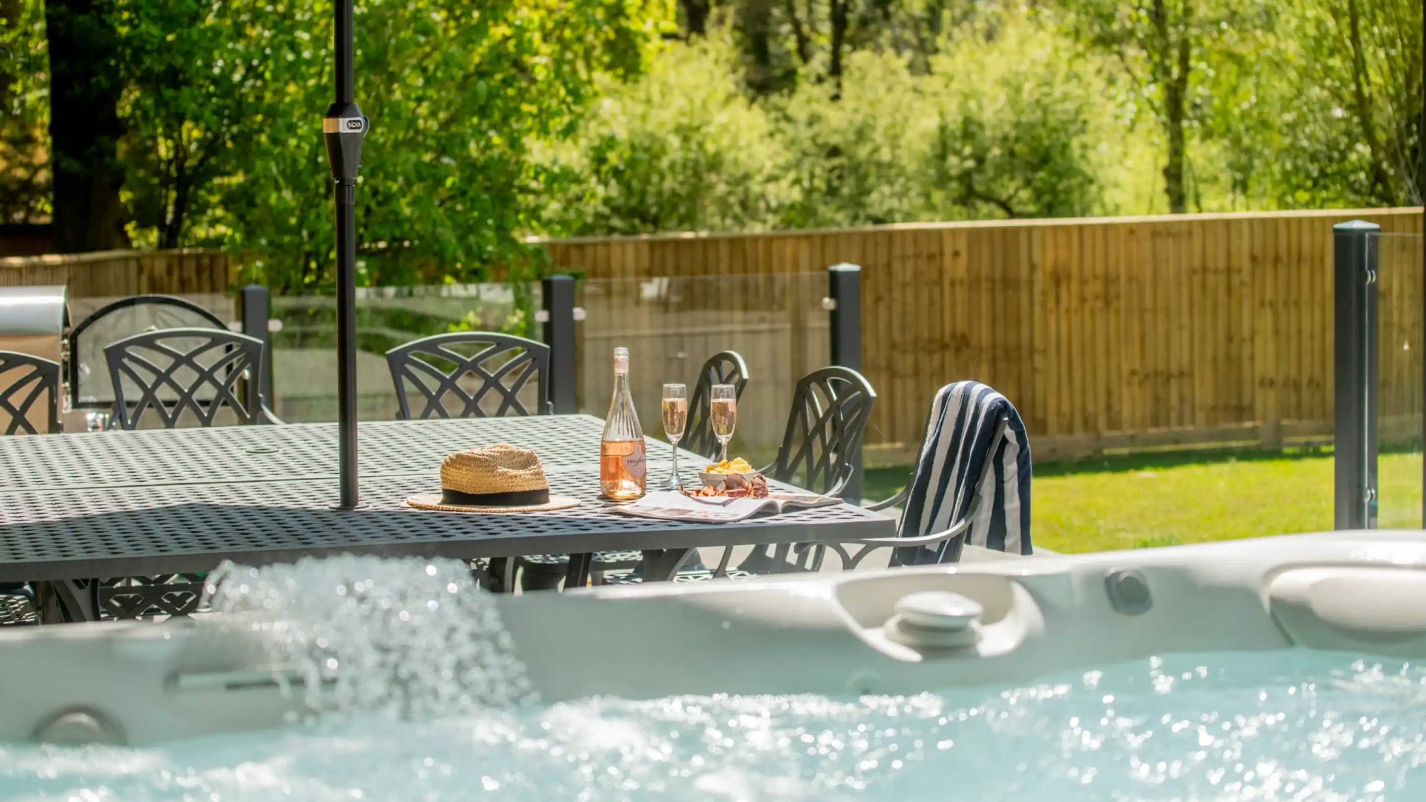 A sunlit outdoor area featuring a hot tub with bubbles, a large dining table with chairs, a bottle of rosé wine, two glasses, a snack plate, and a straw hat. Lush greenery and a wooden fence are visible in the background.