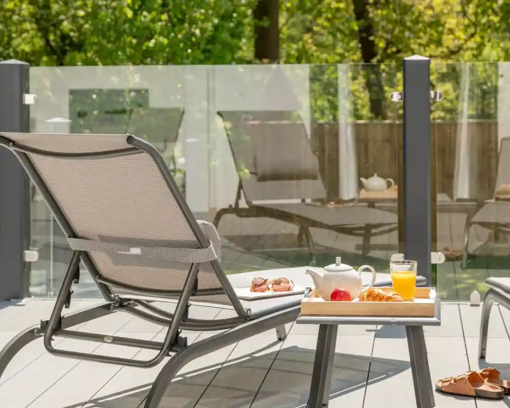 A relaxing outdoor scene featuring a lounge chair next to a tray with a teapot, apple, orange juice, and croissant. The background shows a glass railing and greenery, creating a serene atmosphere.