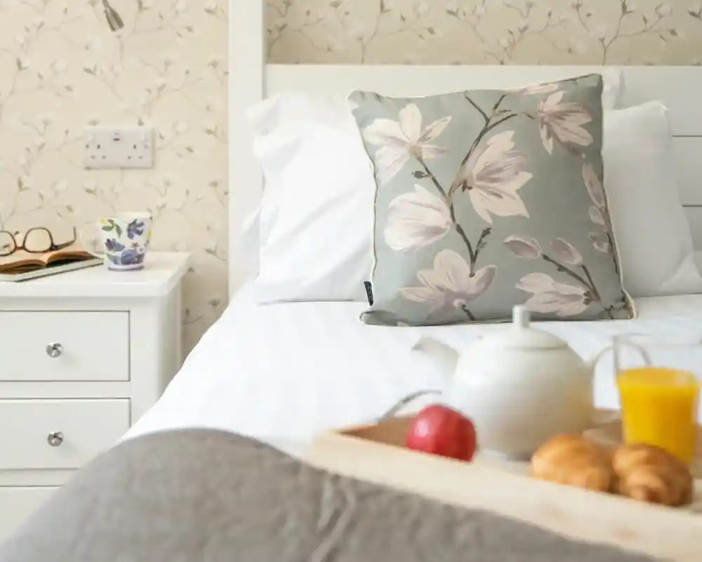 A neatly made bed with a floral-patterned pillow, next to a nightstand holding a cup, a book, and a white teapot on a tray. The tray also has an apple, croissants, and a glass of orange juice. The wall features a subtle floral wallpaper.