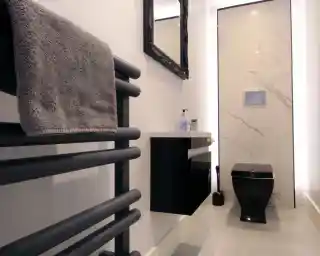 A modern bathroom featuring a black toilet, a sleek wall-mounted vanity with a sink, and a dark towel rail. The wall is decorated with marble tiles, and there's a large mirror above the sink. Soft lighting adds a contemporary feel.