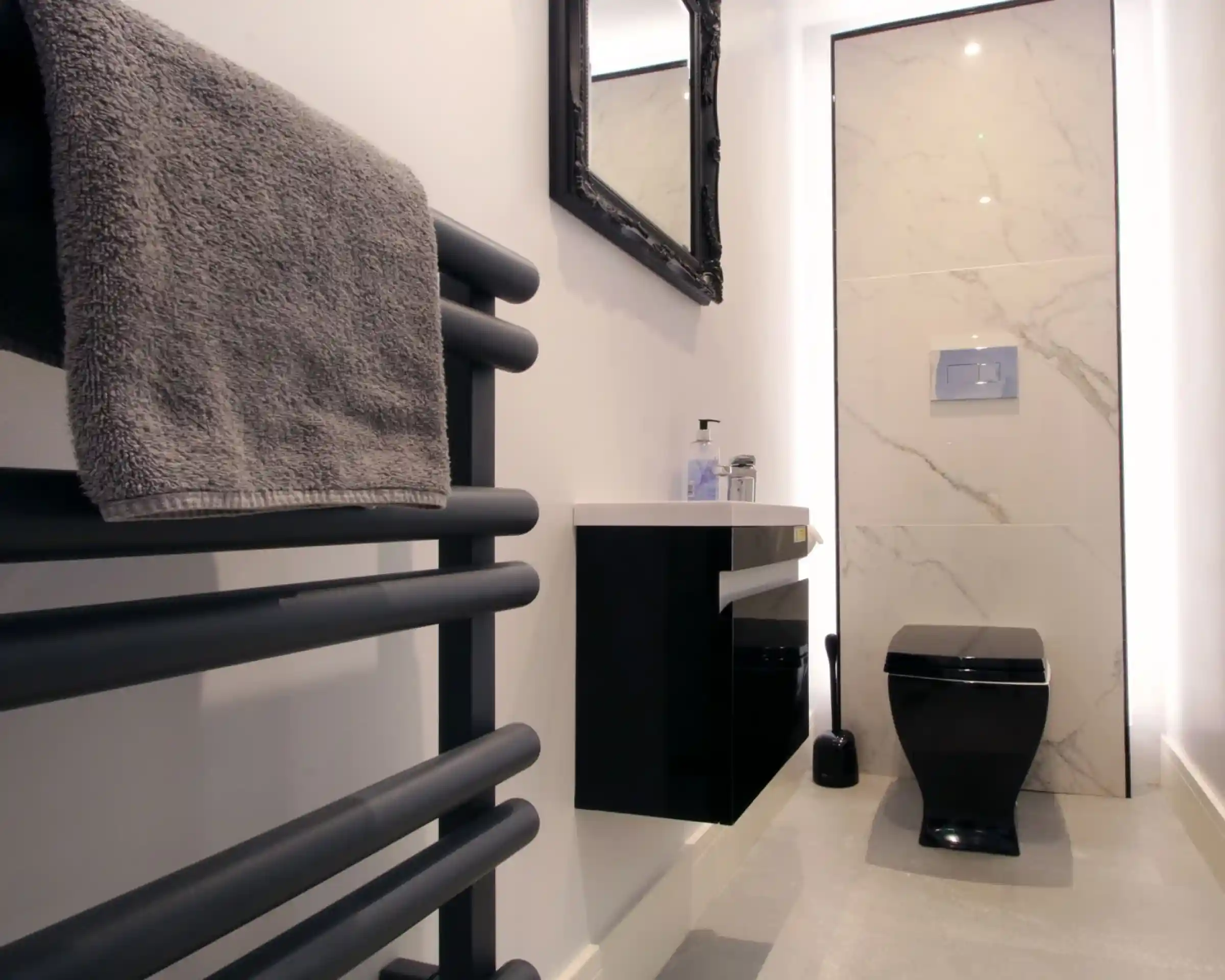 A modern bathroom featuring a black toilet, a sleek wall-mounted vanity with a sink, and a dark towel rail. The wall is decorated with marble tiles, and there's a large mirror above the sink. Soft lighting adds a contemporary feel.