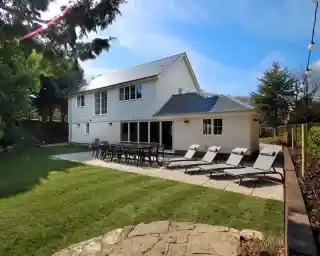 Modern two-story house with a white exterior and large windows, set in a well-maintained garden. Sun loungers are arranged neatly on the patio, accompanied by an outdoor dining area with a table and chairs. Lush green grass surrounds the patio, under a clear blue sky.