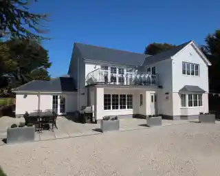 A modern two-story house with a white exterior and dark grey roof. The front features large windows, a balcony, and a patio area with outdoor furniture and planters. A gravel driveway leads up to the entrance.