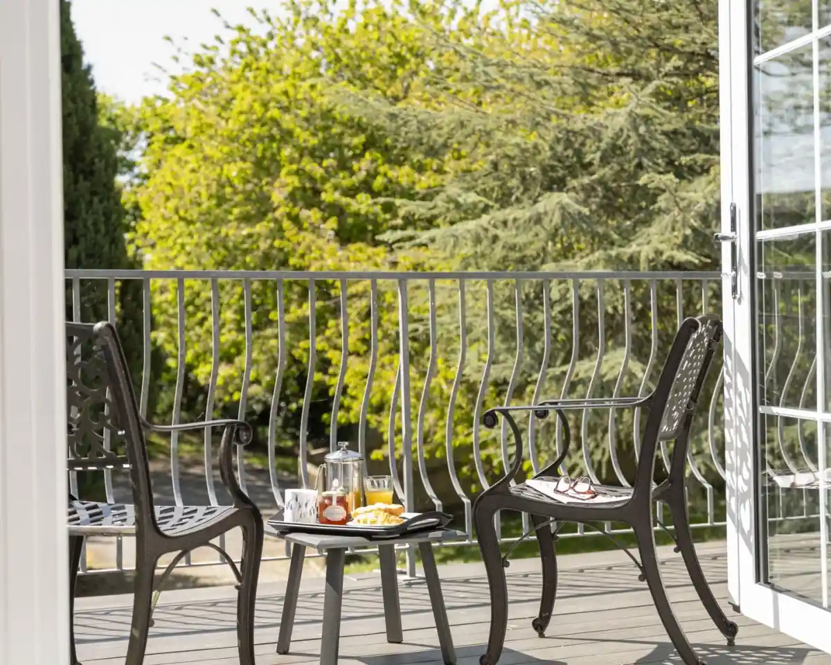 A cozy balcony setup featuring two black metal chairs and a small round table with a French press, pastries, and a glass of orange juice. Lush green trees provide a scenic backdrop.