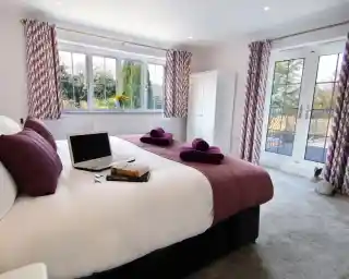 A cozy bedroom featuring two twin beds with purple accents, a laptop on one bed, and neatly arranged towels. Large windows provide natural light and a view of greenery outside. There is a wardrobe and a set of double doors leading to a balcony.