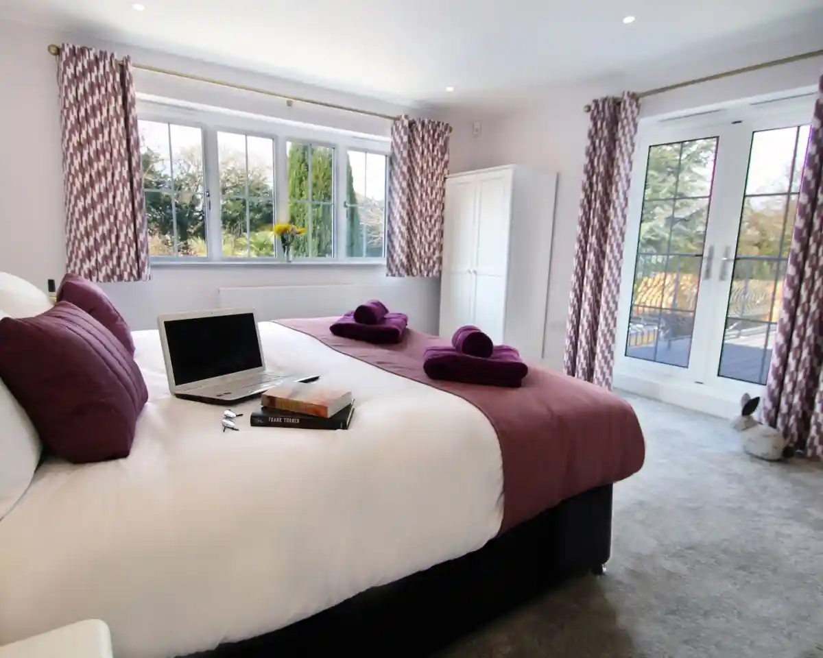 A cozy bedroom featuring two twin beds with purple accents, a laptop on one bed, and neatly arranged towels. Large windows provide natural light and a view of greenery outside. There is a wardrobe and a set of double doors leading to a balcony.
