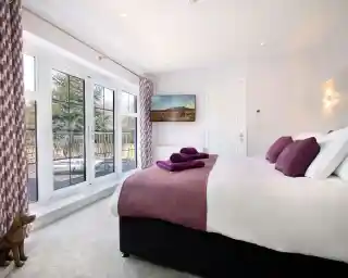 A cozy bedroom featuring a king-size bed with purple accents, two neatly folded towels on the bed, large windows with a view, and a TV mounted on the wall. The decor is modern and inviting, with light-colored walls and plush carpet.