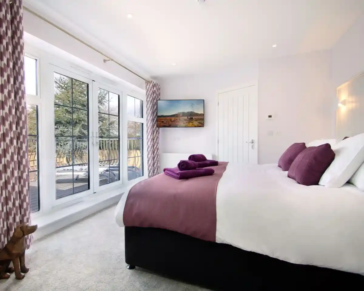 A cozy bedroom featuring a king-size bed with purple accents, two neatly folded towels on the bed, large windows with a view, and a TV mounted on the wall. The decor is modern and inviting, with light-colored walls and plush carpet.