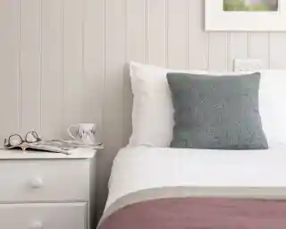 A neatly made bed with a white duvet and a textured gray pillow. Beside the bed, a white bedside table holds a magazine and a decorative mug, with a pair of glasses resting on top.