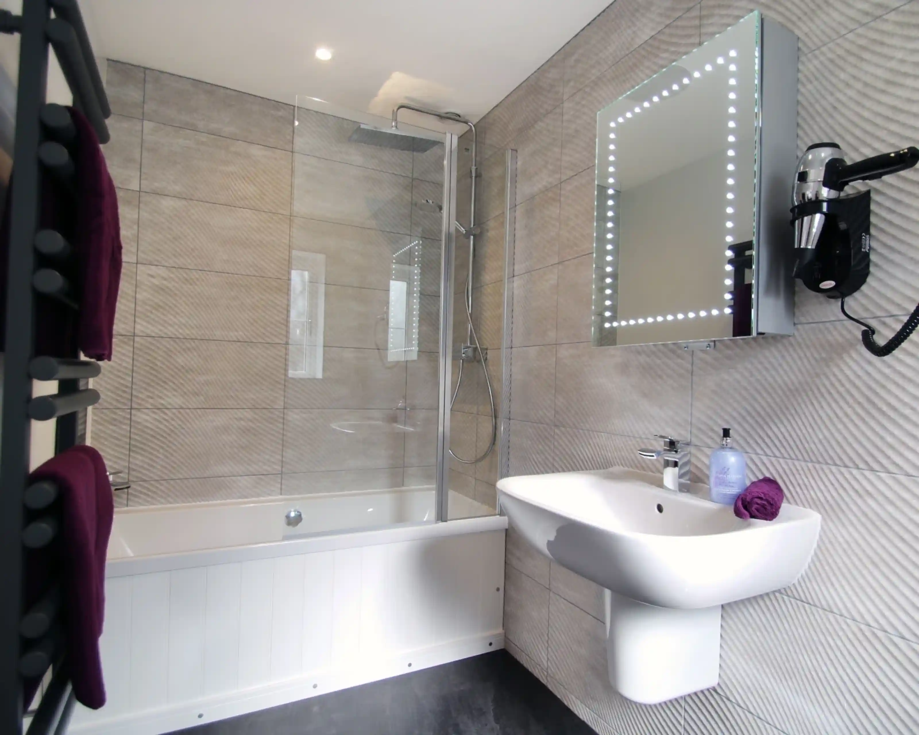 A modern bathroom featuring a glass shower enclosure, a bathtub, a wall-mounted sink, and a illuminated mirror. Towel racks and toiletries are displayed for convenience.