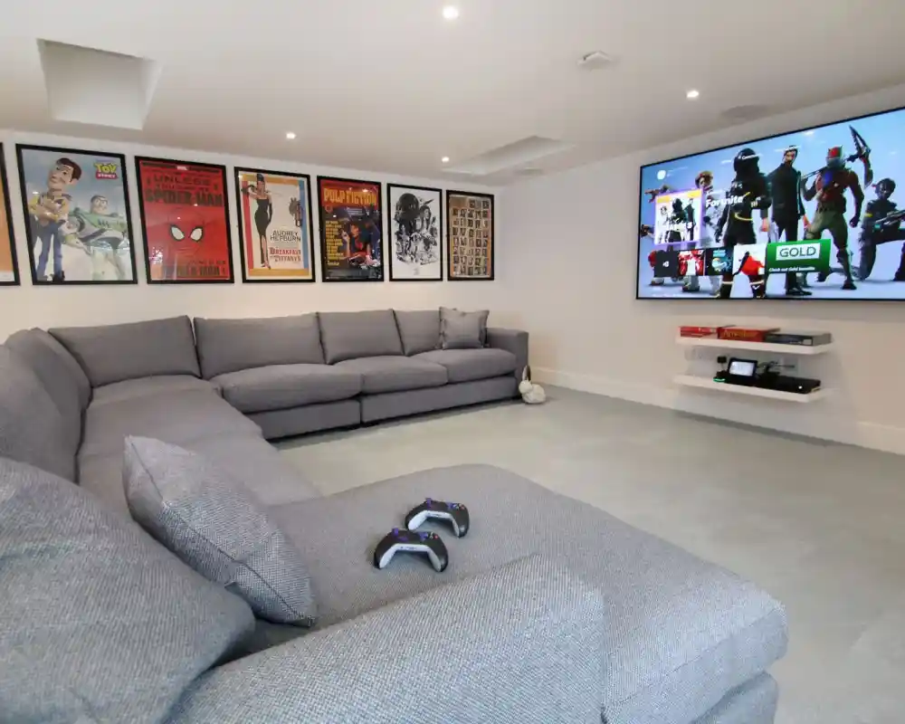 A modern entertainment room featuring a large gray sectional sofa and a wall-mounted TV displaying video game graphics. Various movie posters decorate the walls, adding a vibrant touch to the space. Two gaming controllers are placed on the sofa.