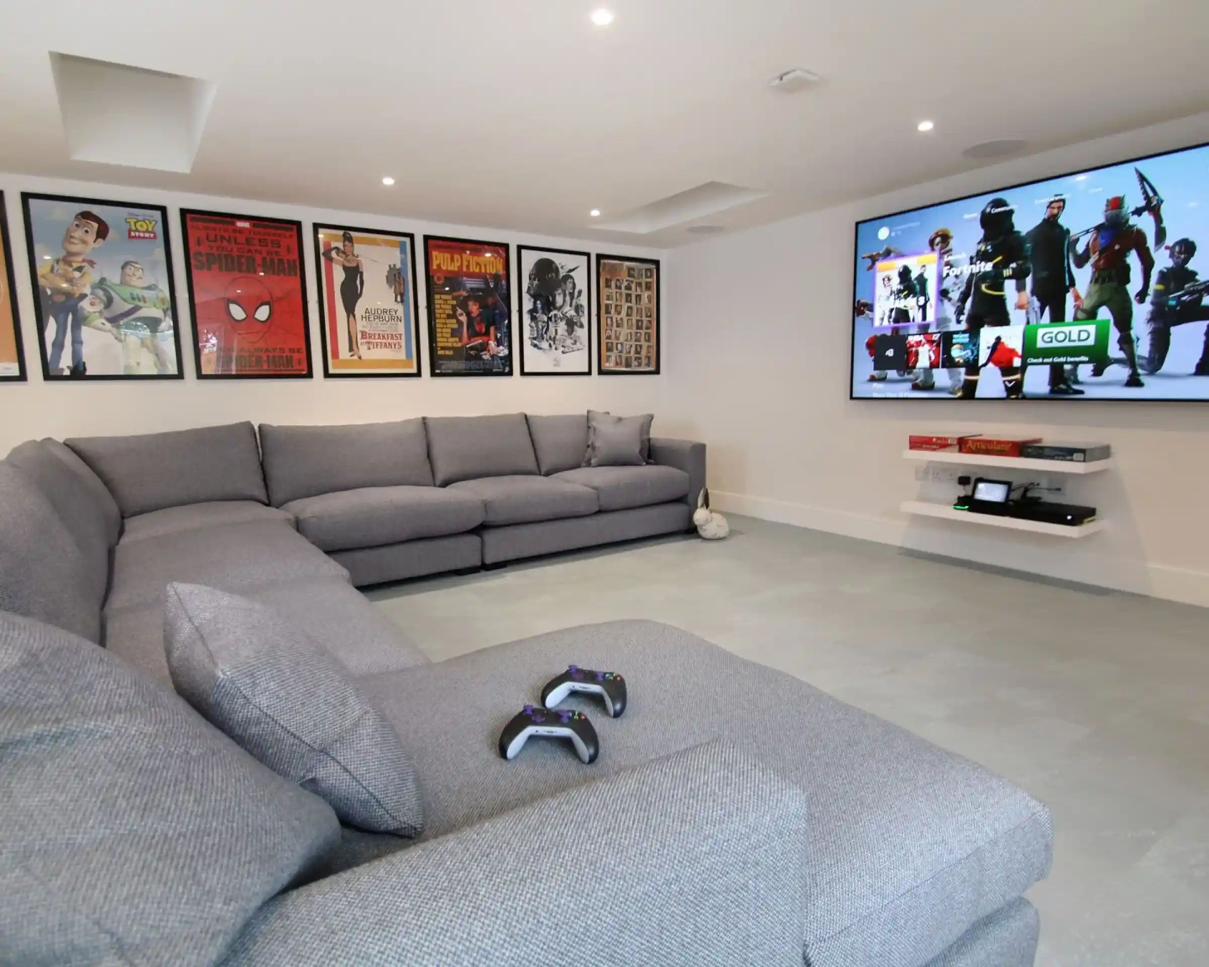 A modern entertainment room featuring a large gray sectional sofa and a wall-mounted TV displaying video game graphics. Various movie posters decorate the walls, adding a vibrant touch to the space. Two gaming controllers are placed on the sofa.