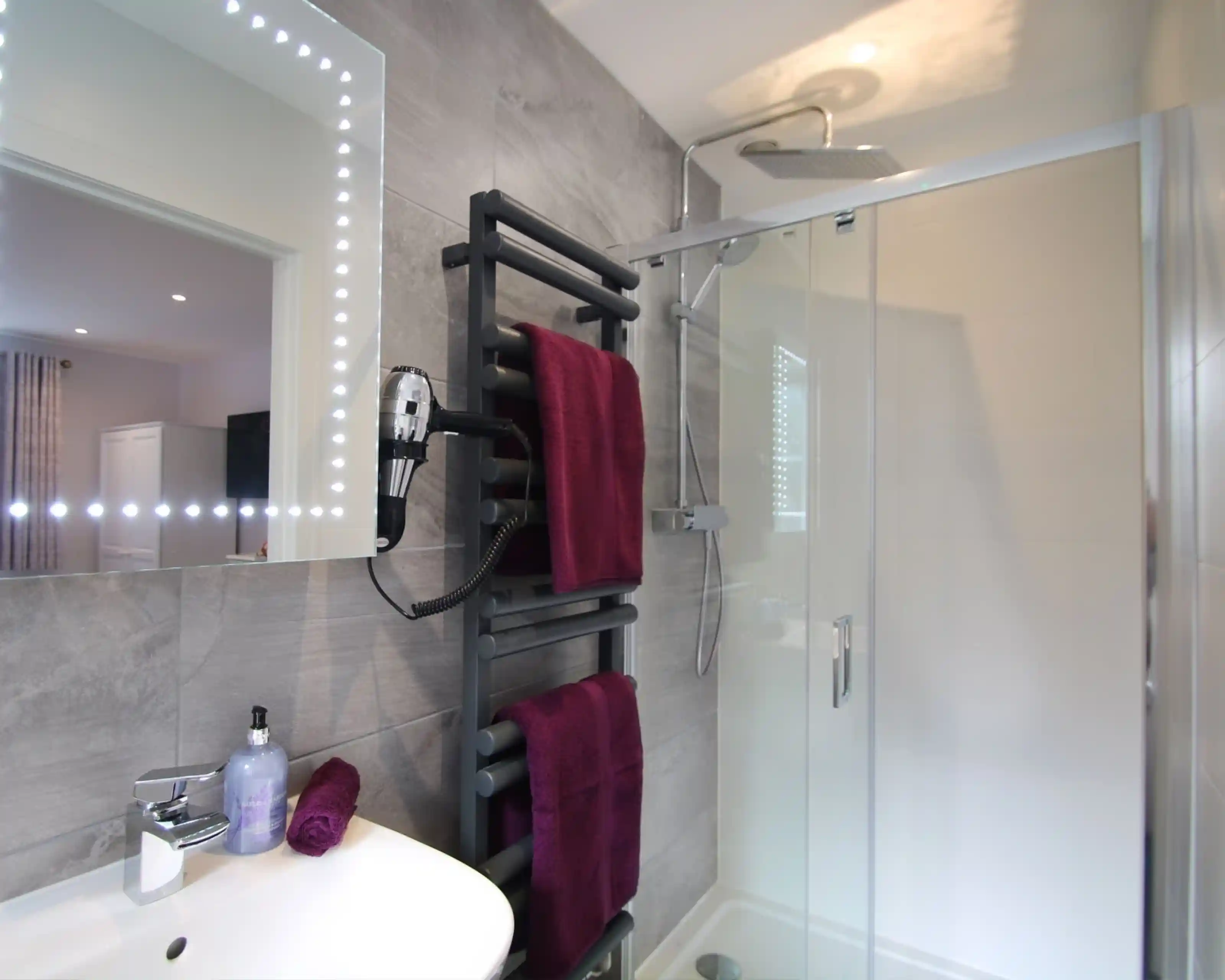 Modern bathroom featuring a glass shower enclosure, a sleek towel warmer with burgundy towels, a stylish sink with a mirror framed by lights, and a contemporary showerhead.