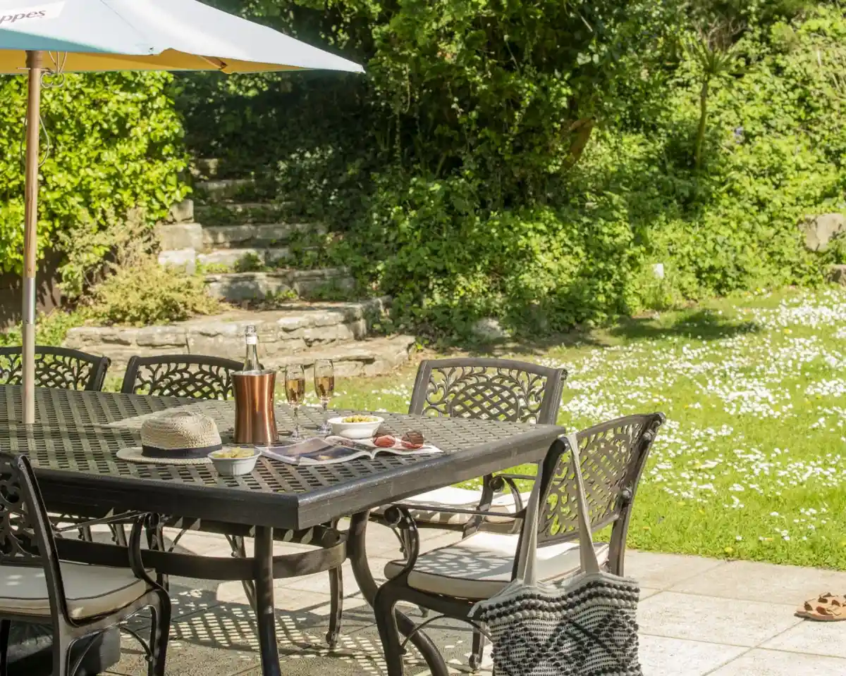 A sunlit patio featuring a dark wrought-iron dining table surrounded by chairs. On the table are glasses, a bottle, and a platter of snacks. An umbrella provides shade, while a grassy area with daisies is visible in the background. Greenery surrounds the scene.