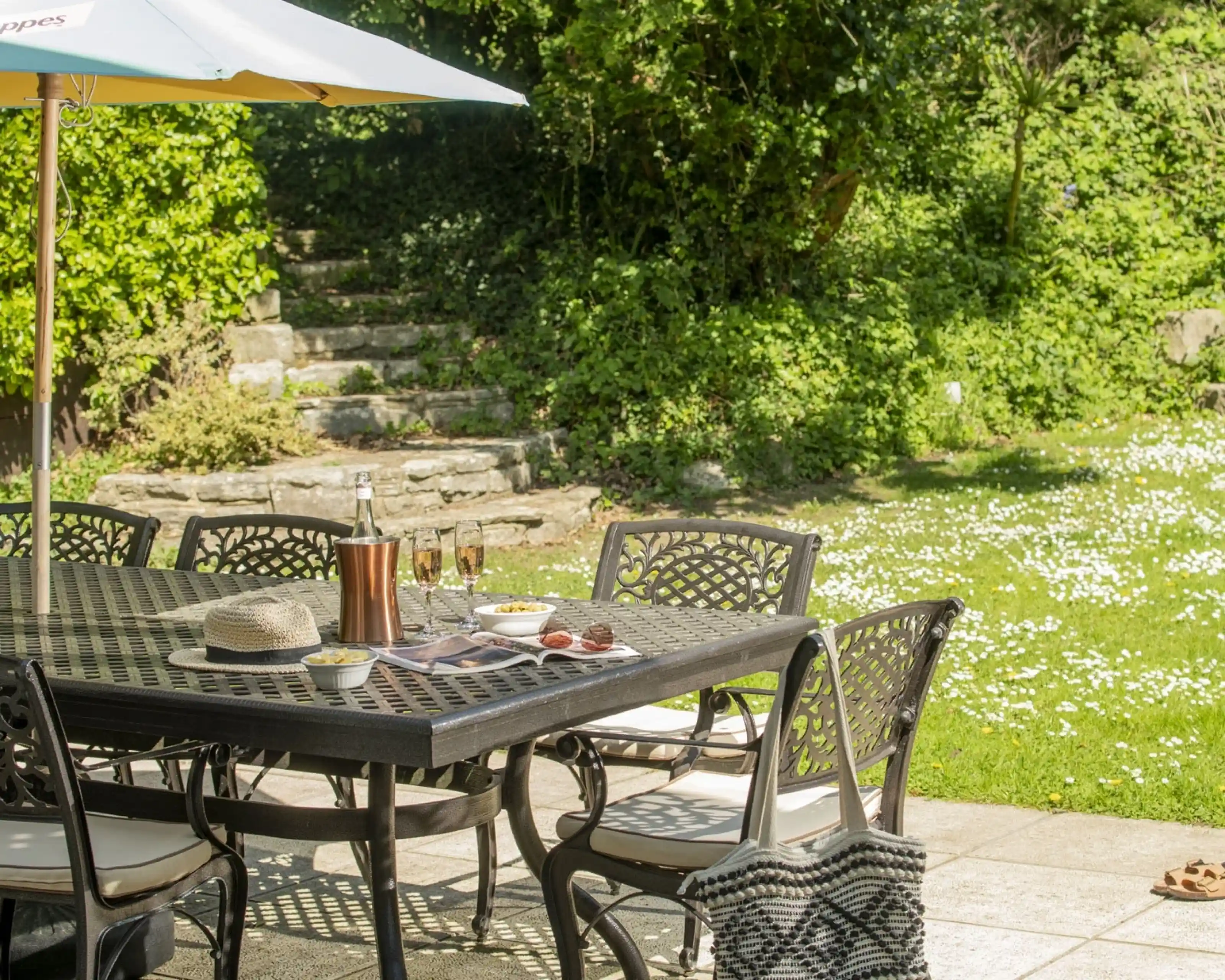 A sunlit patio featuring a dark wrought-iron dining table surrounded by chairs. On the table are glasses, a bottle, and a platter of snacks. An umbrella provides shade, while a grassy area with daisies is visible in the background. Greenery surrounds the scene.