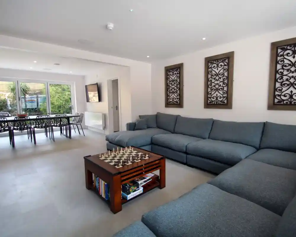 Spacious living room featuring a large blue sectional sofa, a wooden coffee table with a chessboard, and decorative wall panels. The space includes a dining area in the background with a table and chairs, and large windows letting in natural light.