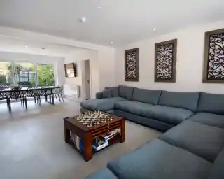 Spacious living room featuring a large blue sectional sofa, a wooden coffee table with a chessboard, and decorative wall panels. The space includes a dining area in the background with a table and chairs, and large windows letting in natural light.