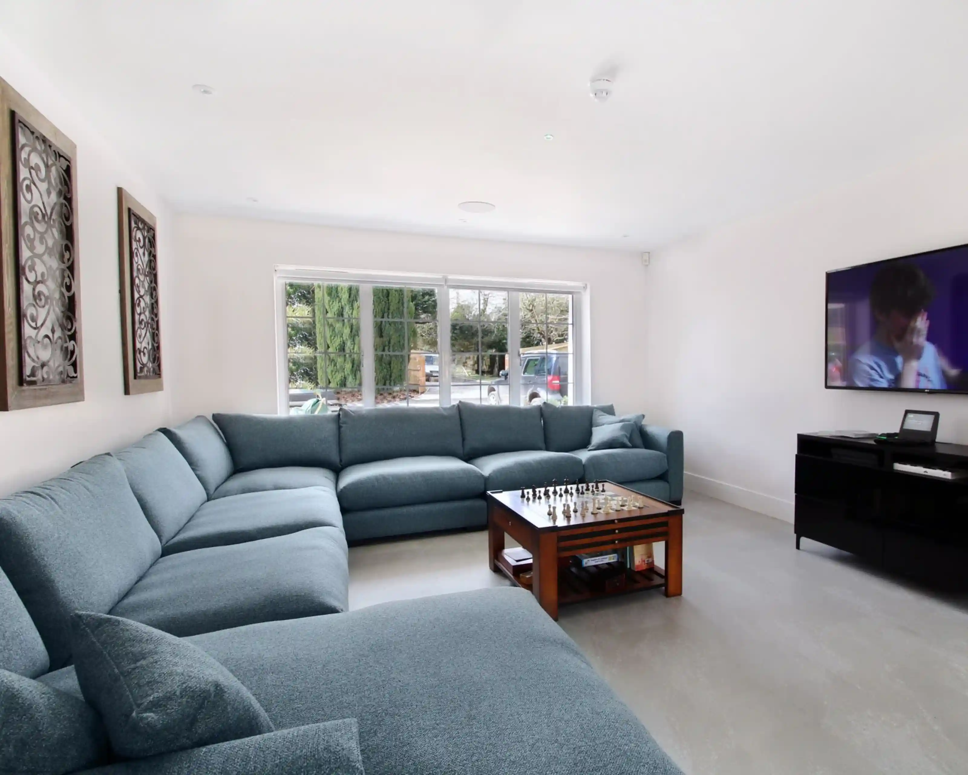 A spacious living room featuring a large, L-shaped blue sectional sofa, a wooden coffee table with a chess set, and a flat-screen TV on a black media console. Large windows provide natural light and a view of greenery outside.