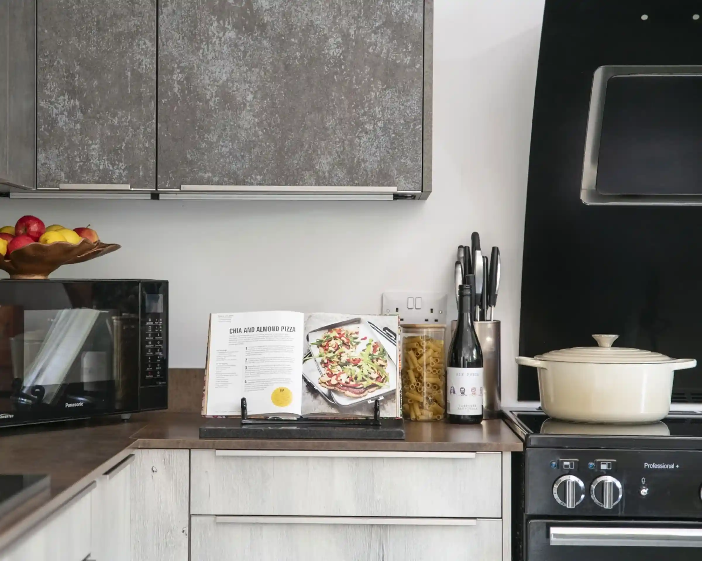 A modern kitchen with a countertop displaying a cookbook open to a pizza recipe. A bowl of fruit sits on the counter, along with a bottle of wine and a jar of pasta. A black stove and microwave are visible in the background.