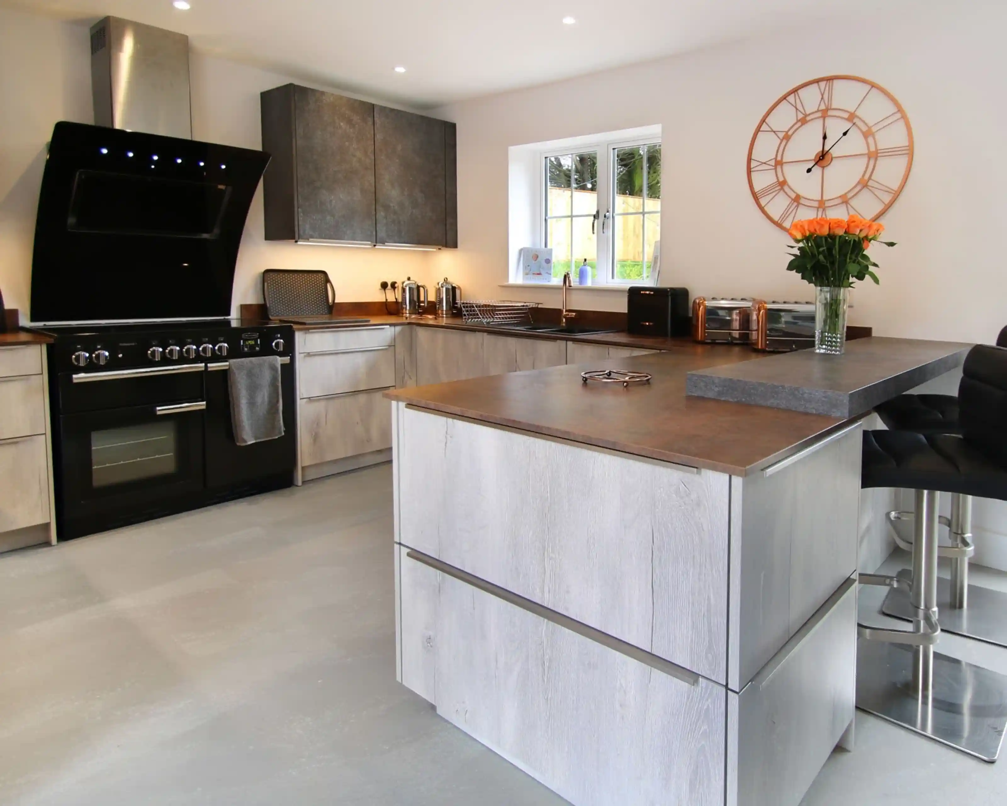 A modern kitchen with a combination of dark and light cabinetry, featuring a black stove, stainless steel appliances, and a large island with a wooden countertop. A vase of orange flowers adds a pop of color on the island, and a stylish wall clock hangs above.