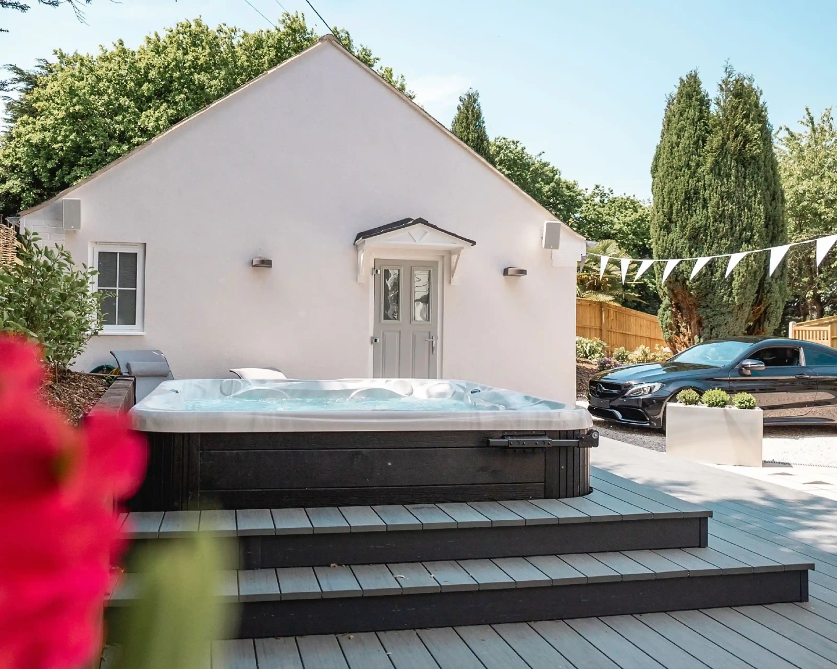A modern outdoor space featuring a hot tub on wooden decking, surrounded by greenery. In the background, a white house with a gray door and windows is visible. A sleek black car is parked nearby, and decorative bunting hangs above.