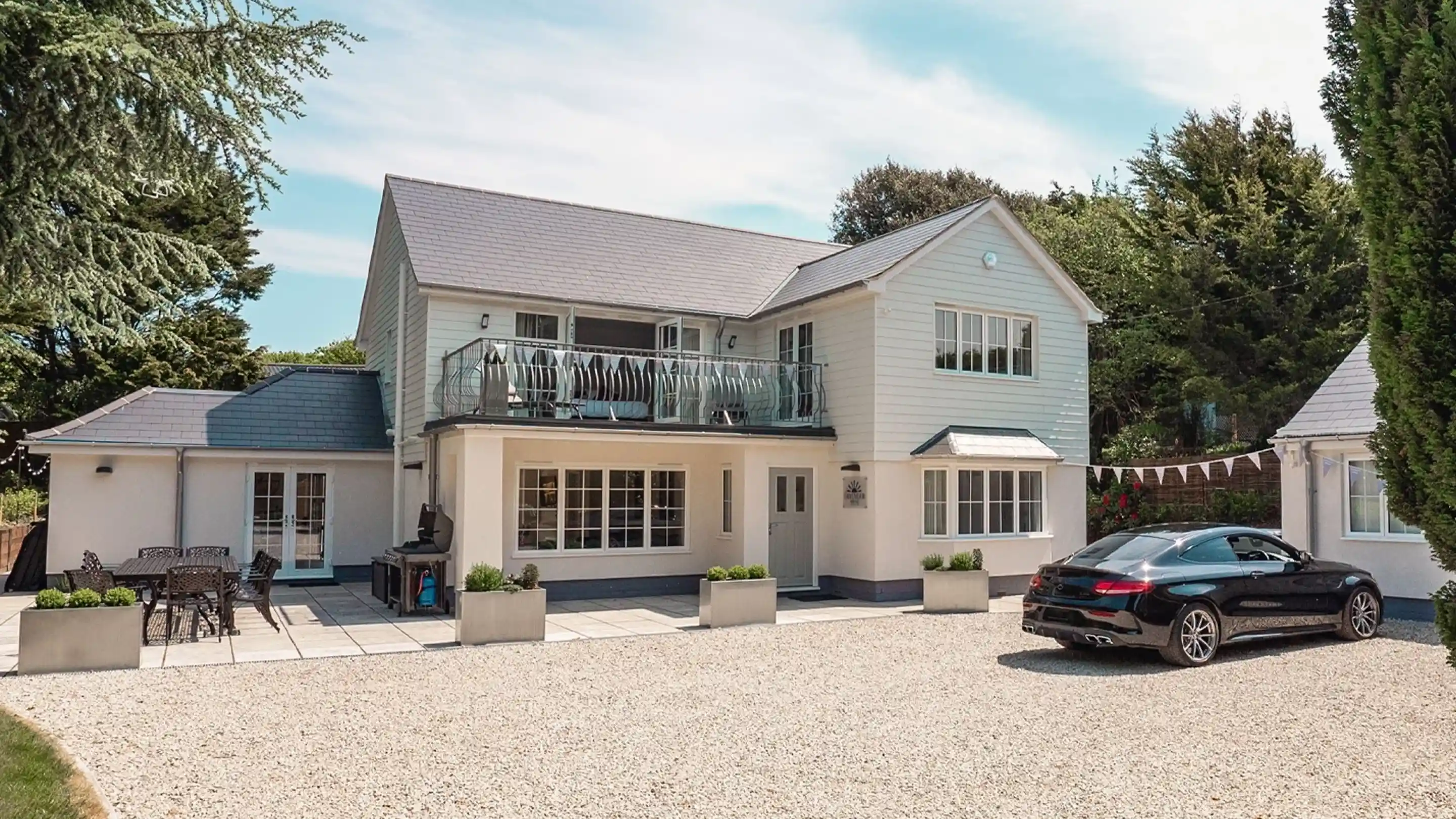 Modern two-story house with a light exterior, featuring a balcony and large windows. A black car is parked on the gravel driveway, and there are outdoor seating arrangements with potted plants. Lush greenery surrounds the property.