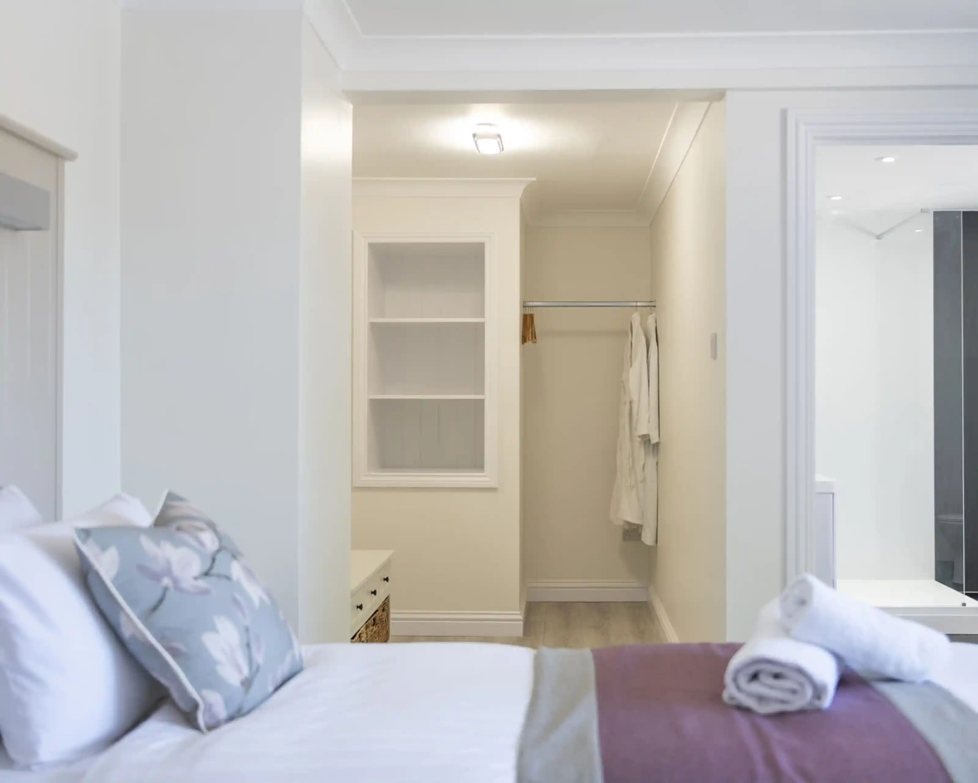 A well-lit bedroom featuring a neatly made bed with decorative pillows. A walk-in closet is partially visible, with a white robe hanging on the wall. The room has soft, neutral-colored walls and a light wood floor.