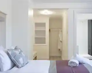 A well-lit bedroom featuring a neatly made bed with decorative pillows. A walk-in closet is partially visible, with a white robe hanging on the wall. The room has soft, neutral-colored walls and a light wood floor.