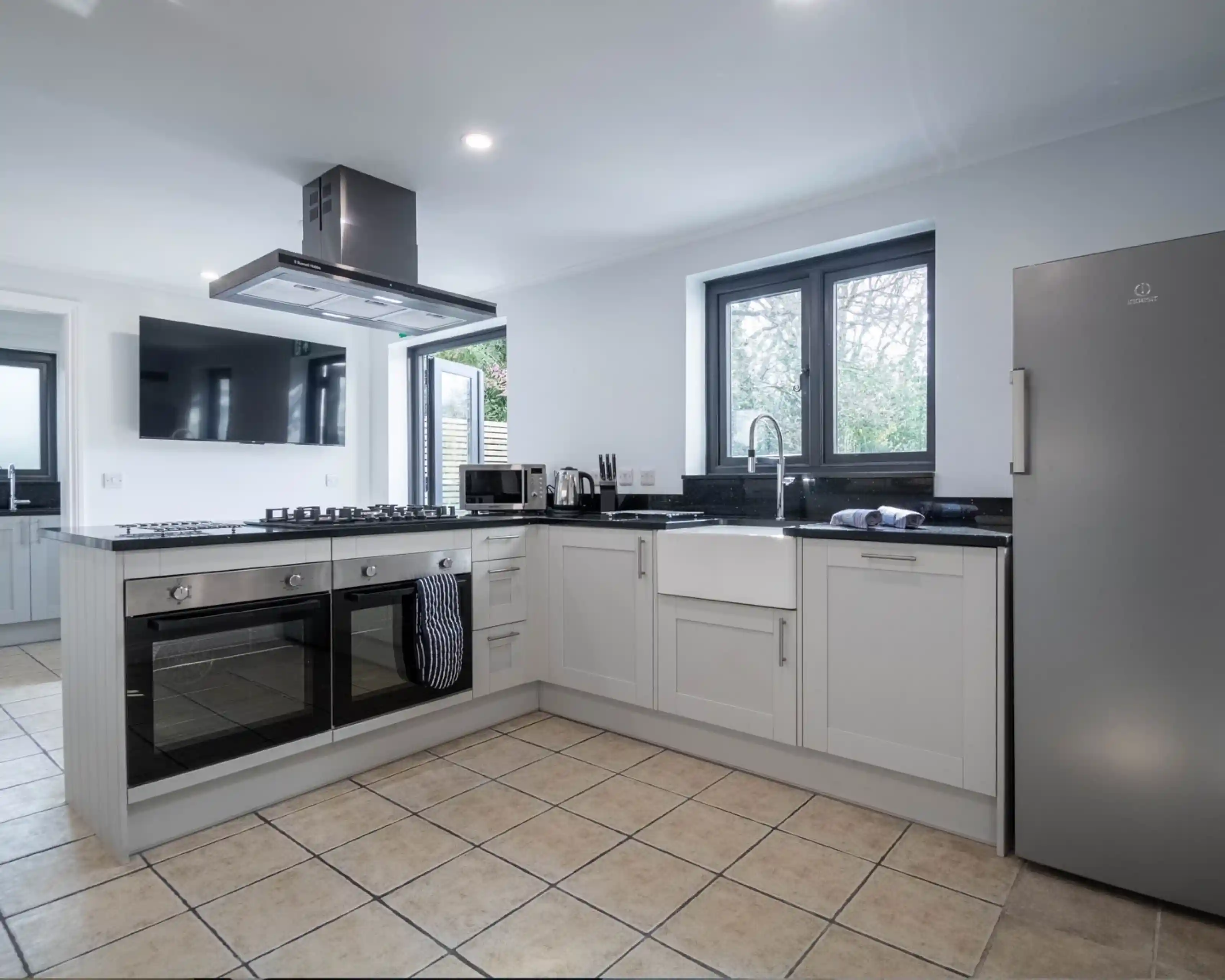 Modern kitchen featuring white cabinetry, a stainless steel oven, and a large window. A gas cooktop with a hood, a farmhouse sink, and tile flooring complete the space.
