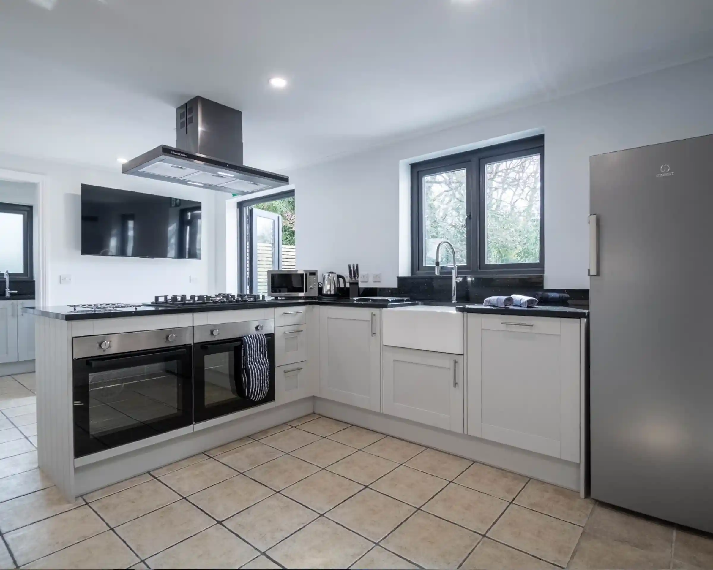 Modern kitchen featuring white cabinetry, a stainless steel oven, and a large window. A gas cooktop with a hood, a farmhouse sink, and tile flooring complete the space.