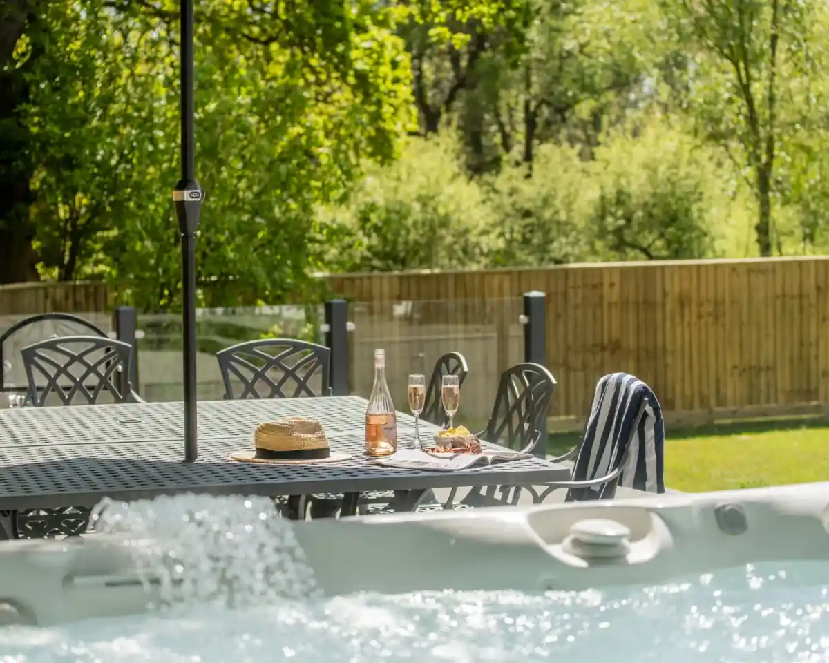 A sunny outdoor space featuring a table set with two glasses of champagne and a bottle of rosé. A straw hat and a light blue striped towel are on the table. In the foreground, a hot tub bubbles, with greenery and a wooden fence in the background.