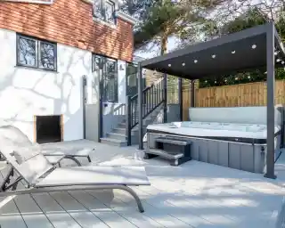 A backyard patio features a hot tub under a sleek awning, accompanied by a lounge chair. There are steps leading to an interior door, with a wooden fence and trees in the background.