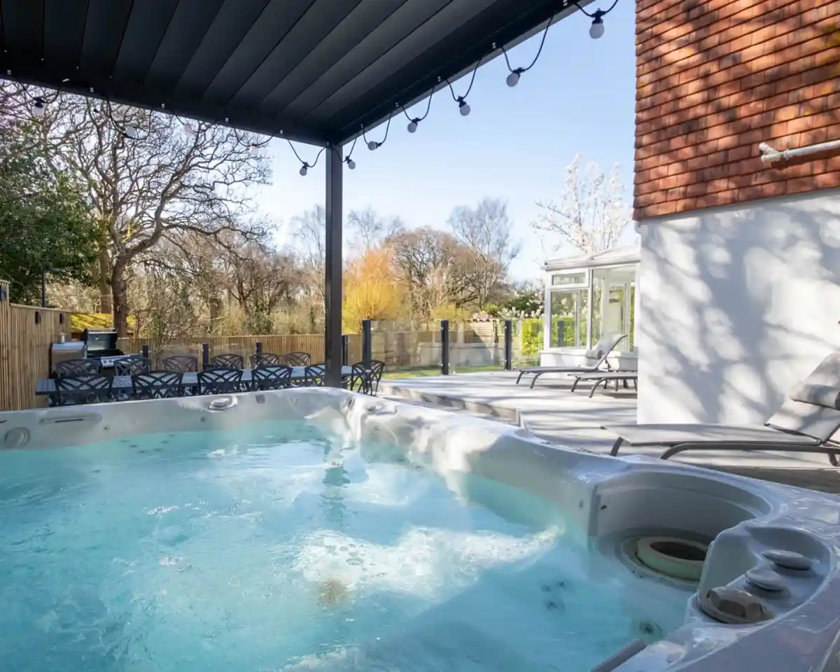 A hot tub filled with water sits under a canopy; nearby are lounge chairs and a glimpsed garden with trees in the background.