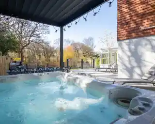 A hot tub filled with water sits under a canopy; nearby are lounge chairs and a glimpsed garden with trees in the background.