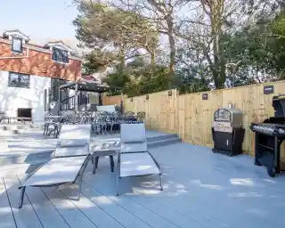 A modern outdoor patio featuring wooden decking, two gray lounge chairs, a barbecue grill, and a dining area with black chairs. The background includes a house surrounded by trees.