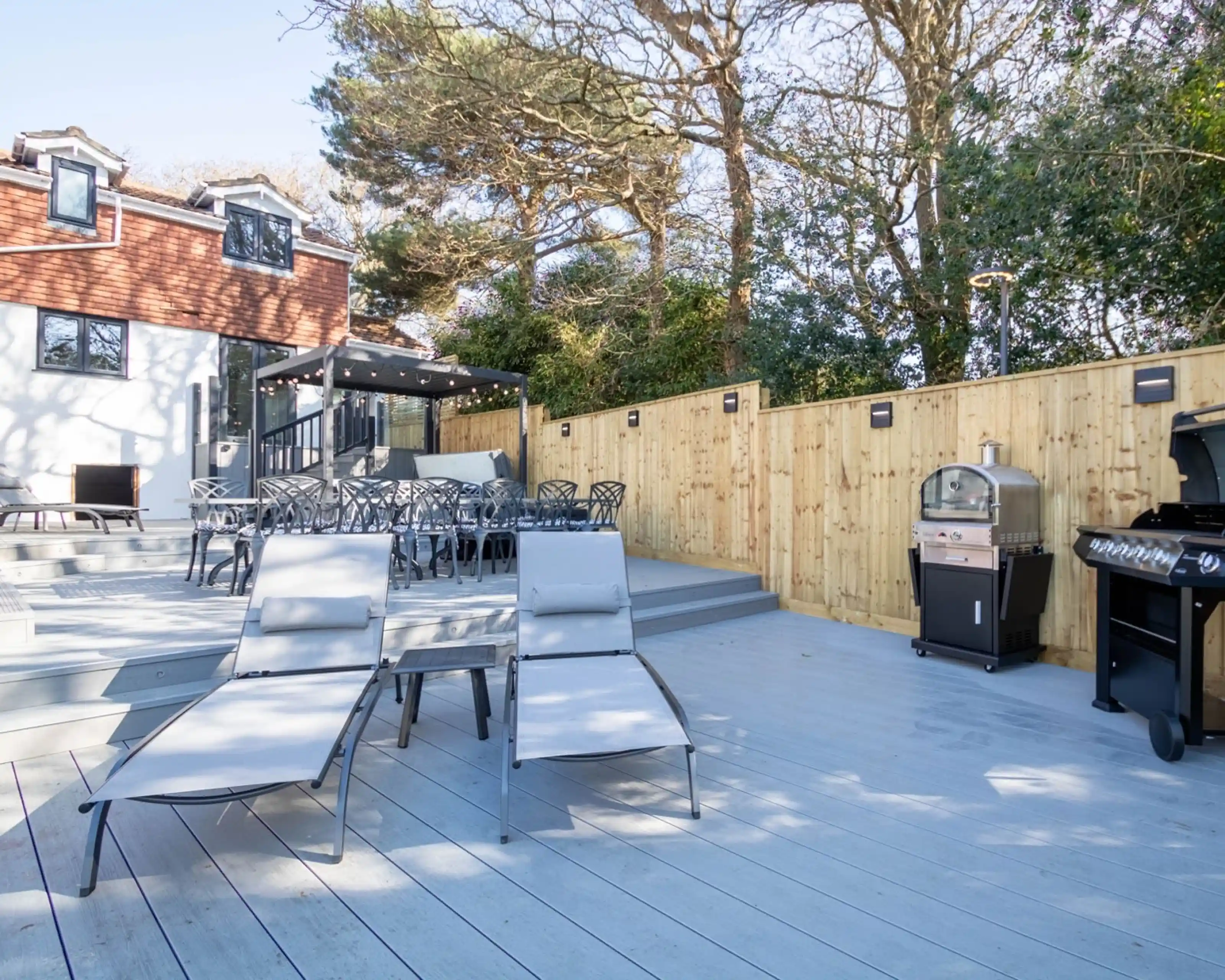 A modern outdoor patio featuring wooden decking, two gray lounge chairs, a barbecue grill, and a dining area with black chairs. The background includes a house surrounded by trees.