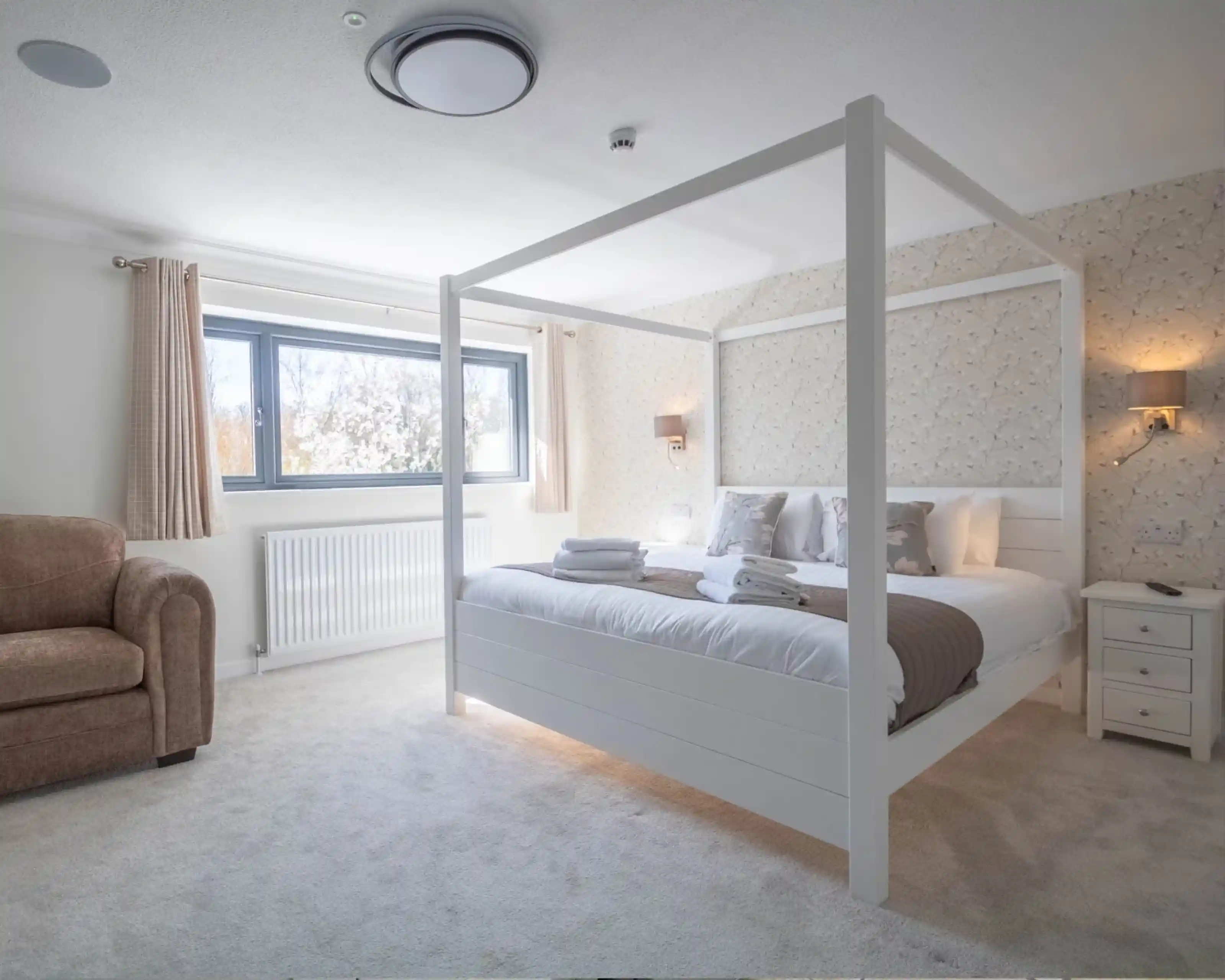 Bright and cozy bedroom featuring a four-poster bed with white frame, light bedding, and decorative pillows. A plush armchair sits in one corner, and the room has soft, natural light coming through a window. Stylish wall coverings and light fixtures add a modern touch.