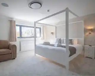 Bright and cozy bedroom featuring a four-poster bed with white frame, light bedding, and decorative pillows. A plush armchair sits in one corner, and the room has soft, natural light coming through a window. Stylish wall coverings and light fixtures add a modern touch.