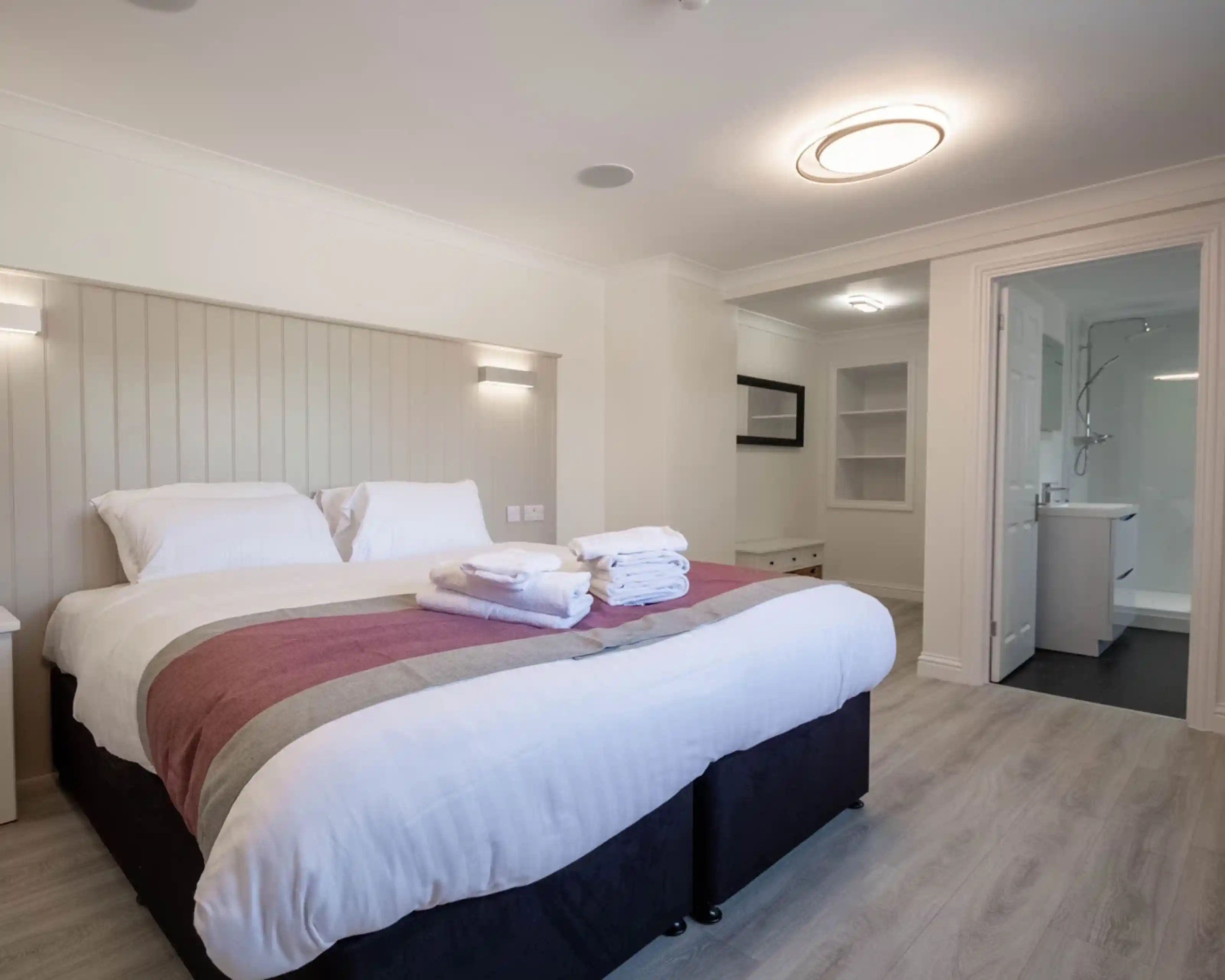 A modern hotel room featuring a neatly made bed with white linens and a maroon throw blanket, accompanied by stacked towels. The room has light wood flooring and a minimalistic design, with a door leading to a bathroom visible in the background.