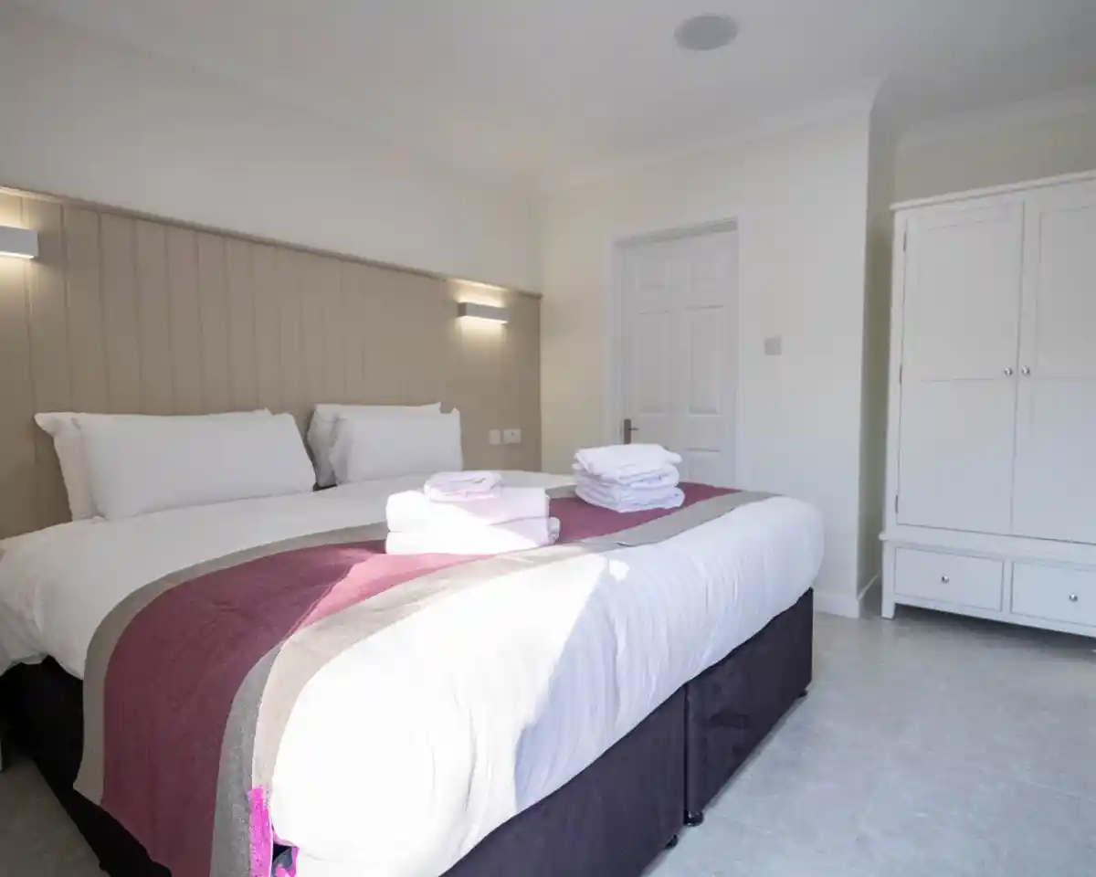 A well-lit bedroom featuring two twin beds with white linens and a maroon throw blanket. Neatly stacked towels rest on the bed. A wardrobe is visible in the background against a light-colored wall.
