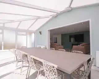 A spacious dining area with a large wooden table surrounded by intricately designed chairs. The room features large glass windows and a light-colored wall, creating a bright and airy atmosphere. In the background, a cozy living area is visible with two sofas and a television.