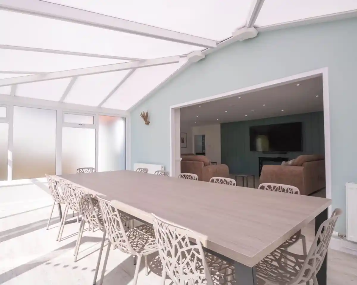 A spacious dining area with a large wooden table surrounded by intricately designed chairs. The room features large glass windows and a light-colored wall, creating a bright and airy atmosphere. In the background, a cozy living area is visible with two sofas and a television.