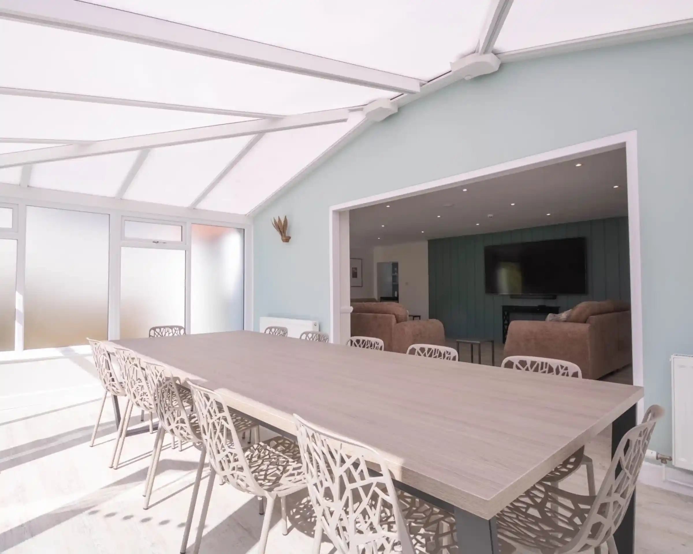 A spacious dining area with a large wooden table surrounded by intricately designed chairs. The room features large glass windows and a light-colored wall, creating a bright and airy atmosphere. In the background, a cozy living area is visible with two sofas and a television.