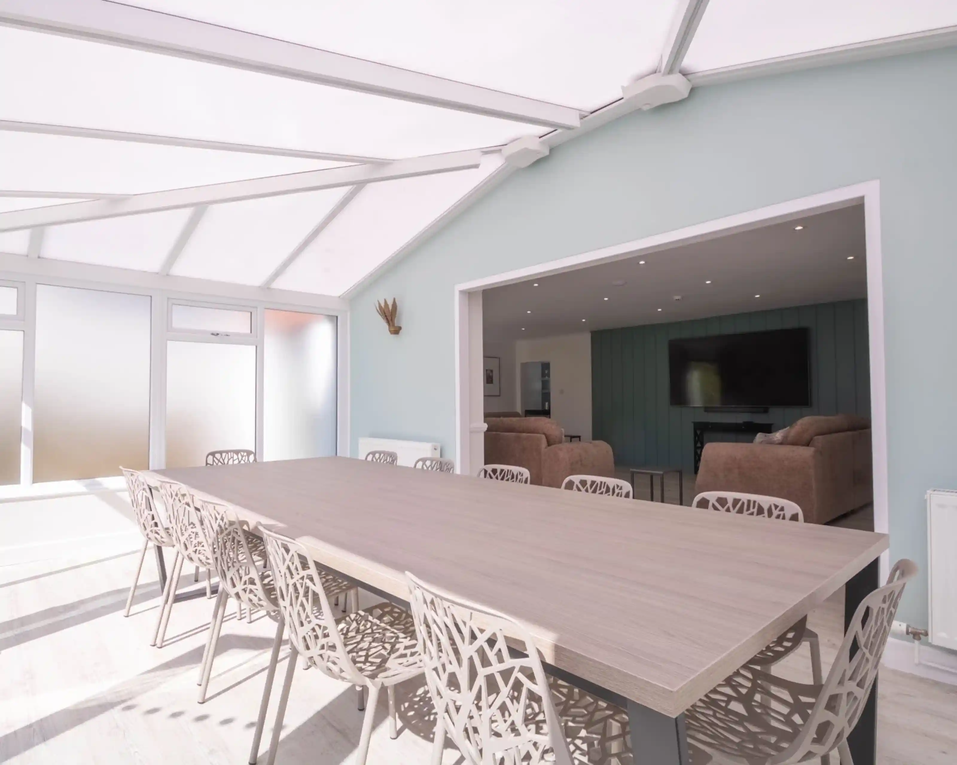 A spacious dining area with a large wooden table surrounded by intricately designed chairs. The room features large glass windows and a light-colored wall, creating a bright and airy atmosphere. In the background, a cozy living area is visible with two sofas and a television.