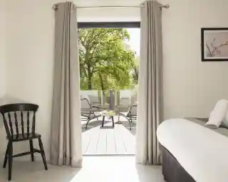 A modern room with a black chair, gray curtains, and a bed, featuring large sliding doors that open to a sunny terrace with lounging chairs and a table. A green tree is visible outside.