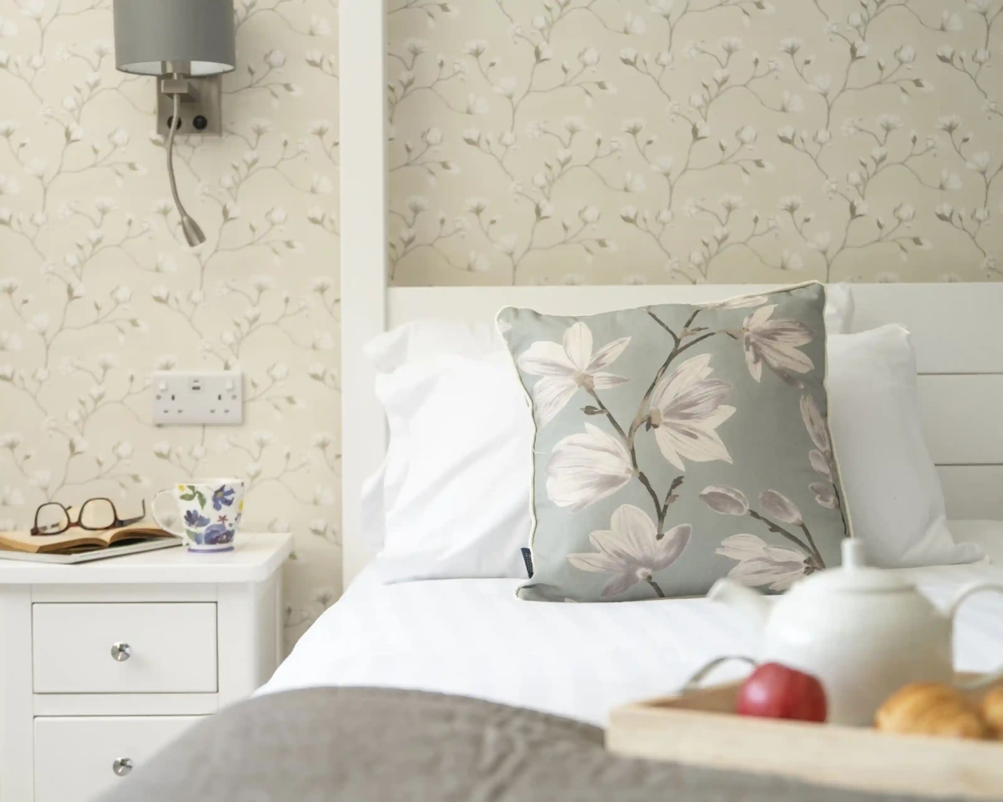 A cozy bedroom with a white bed featuring a decorative floral pillow. A bedside table holds a cup, glasses, and a small wooden tray with an apple and pastries. The wall has a light floral wallpaper.